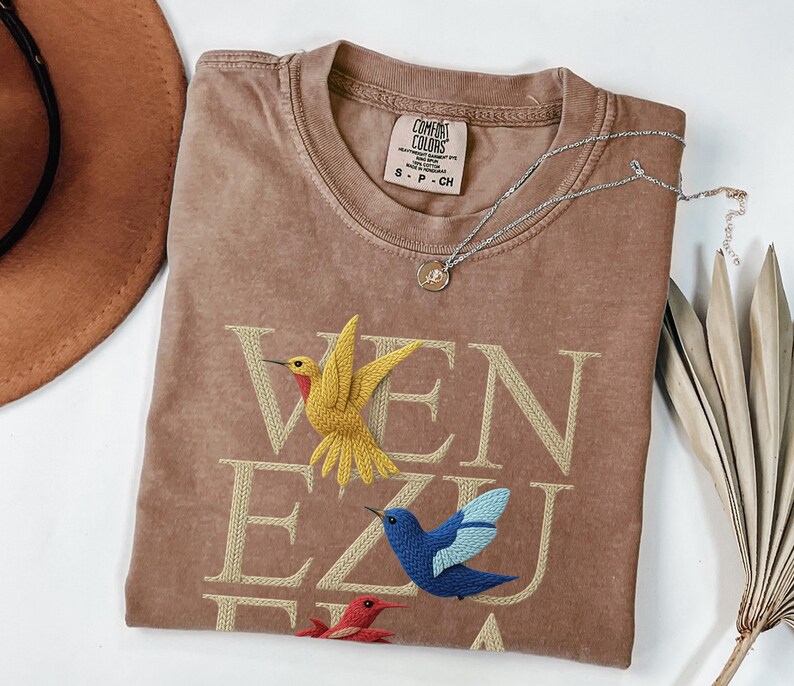 May include: A brown t-shirt with the word "VENEZUELA" embroidered in a light tan colour. The shirt features embroidered hummingbirds in yellow, blue, and red. A silver necklace with a small pendant rests on the shirt.