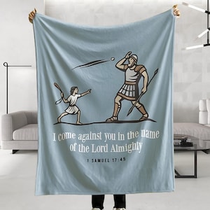 May include: Light blue blanket featuring a graphic illustration of two figures, one throwing a stone at the other. The text "I come against you in the name of the Lord Almighty" is printed below the figures. The blanket is held up by a person.