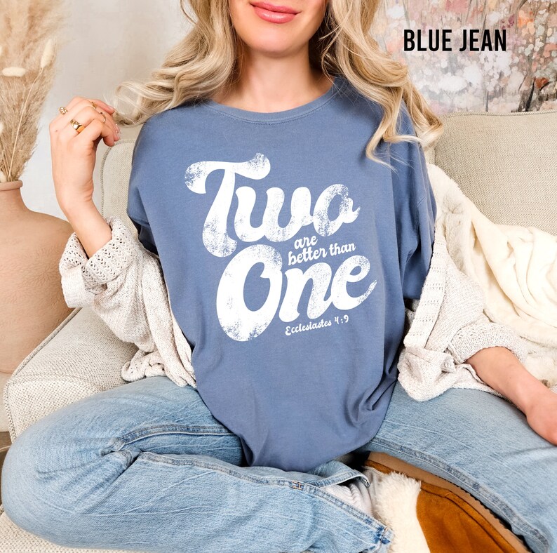 May include: A blue jean colored t-shirt featuring white distressed lettering that reads Two are better than One and Ecclesiastes 4:9. The shirt has a crew neckline and short sleeves, showcasing a vintage-inspired graphic print on the front chest.