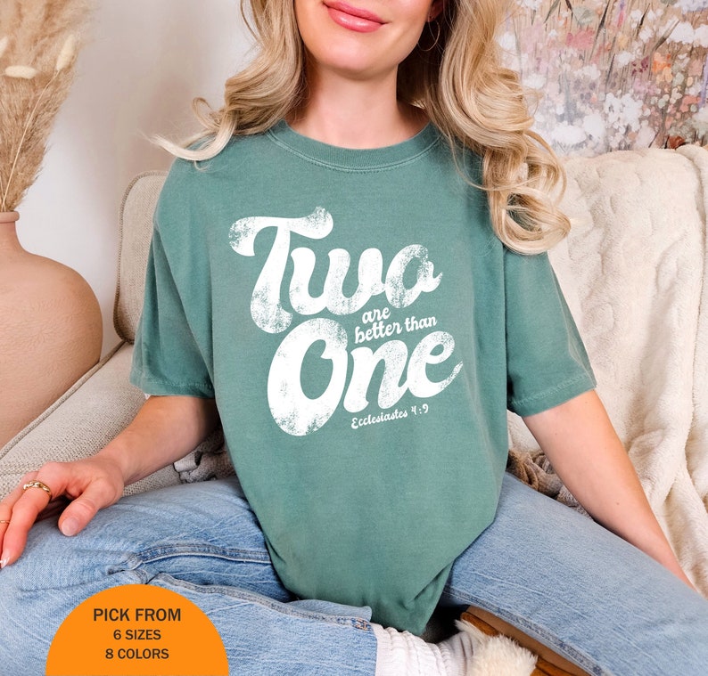 May include: A sage green short-sleeve t-shirt featuring white distressed lettering that reads Two are better than One and Ecclesiastes 4:9. The shirt has a crew neckline and a relaxed fit, with an orange circular graphic at the bottom left corner.
