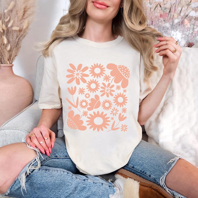 May include: Cream-colored t-shirt featuring a peach-colored floral design. The design includes various flower shapes and sizes, creating a cohesive pattern. The shirt is made of a soft material and is suitable for casual wear.