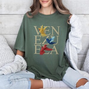 May include: An olive green t-shirt featuring the word "VENEZUELA" in a stacked design. The letters are in a tan colour, and three colourful birds are placed above the letters. The birds are yellow, blue, and red.