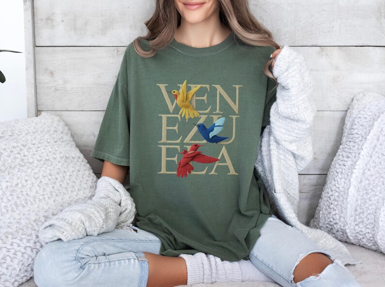 May include: An olive green t-shirt featuring the word "VENEZUELA" in a stacked design. The letters are in a tan colour, and three colourful birds are placed above the letters. The birds are yellow, blue, and red.