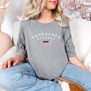 Pode incluir: Uma camiseta cinza com a palavra "VENEZUELA" impressa em branco com um pequeno gr&aacute;fico da bandeira venezuelana abaixo. A camiseta est&aacute; sendo usada por uma pessoa sentada em um sof&aacute; branco.