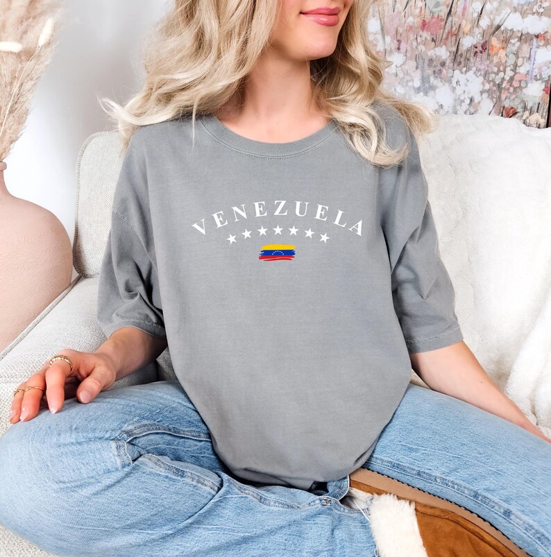 Pode incluir: Uma camiseta cinza com a palavra "VENEZUELA" impressa em branco com um pequeno gr&aacute;fico da bandeira venezuelana abaixo. A camiseta est&aacute; sendo usada por uma pessoa sentada em um sof&aacute; branco.