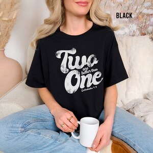 May include: A black short-sleeved t-shirt featuring white distressed-style text that reads Two are better than One and Ecclesiastes 4:9. The shirt has a crew neckline and a relaxed fit, with the word BLACK printed in the upper right corner.