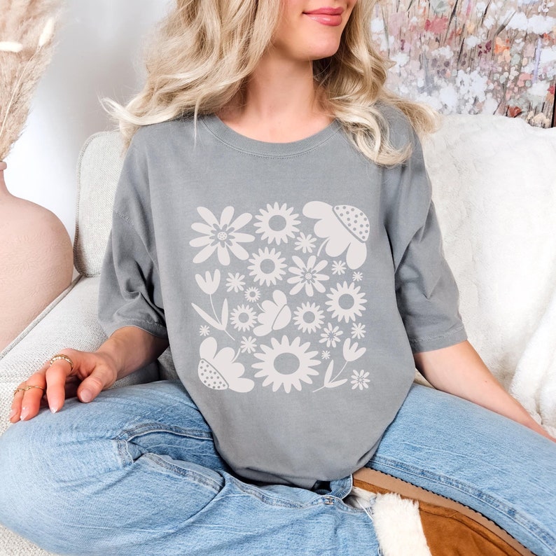May include: A gray t-shirt featuring a floral design in a square arrangement. The design includes various flower shapes and a butterfly. The shirt has short sleeves and a crew neck.