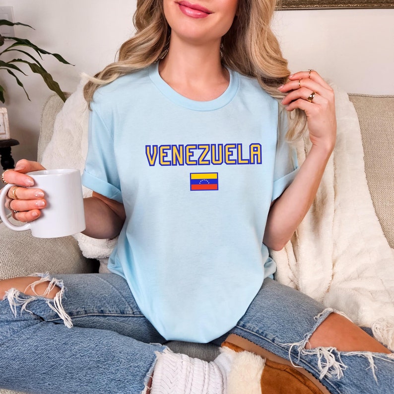 May include: Light blue t-shirt with the word "VENEZUELA" in yellow and blue lettering, and a small Venezuelan flag graphic. The shirt is made of a soft material.