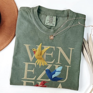 May include: Olive green t-shirt with embroidered hummingbirds and the word "VENEZUELA" in tan thread. A gold necklace with a small pendant rests on the shirt. The shirt is folded on a white surface.