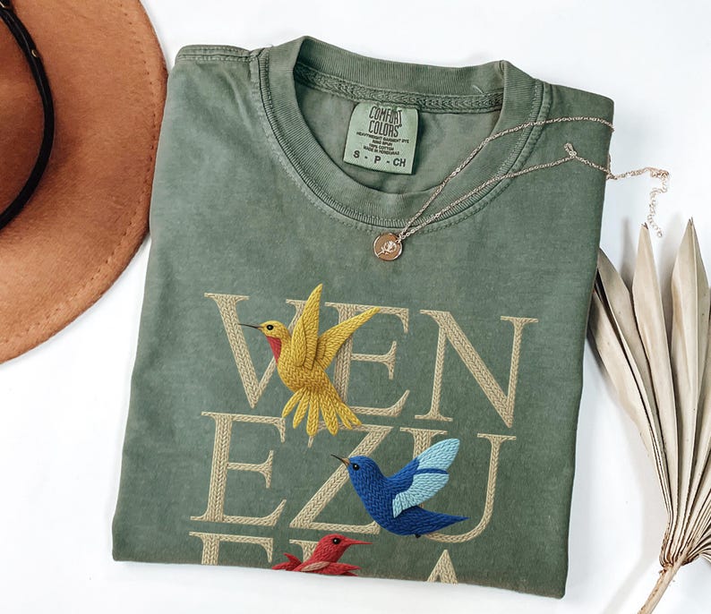 May include: Olive green t-shirt with embroidered hummingbirds and the word "VENEZUELA" in tan thread. A gold necklace with a small pendant rests on the shirt. The shirt is folded on a white surface.