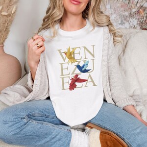 May include: White crew neck t-shirt featuring the word "VENEZUELA" in gold, with illustrations of yellow, blue, and red birds. The shirt is a casual style.