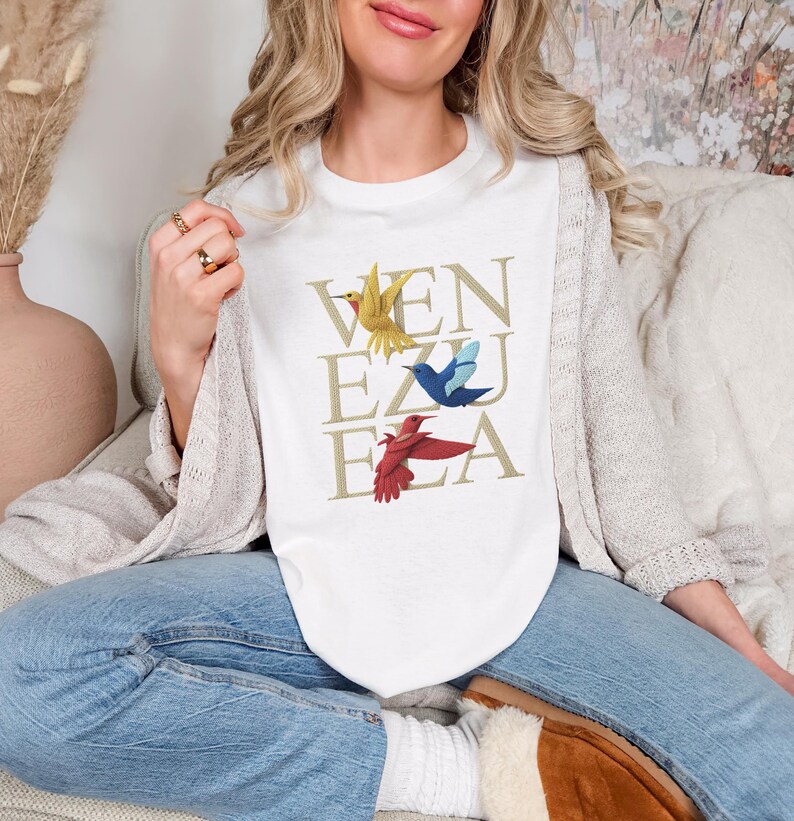 May include: White crew neck t-shirt featuring the word "VENEZUELA" in gold, with illustrations of yellow, blue, and red birds. The shirt is a casual style.