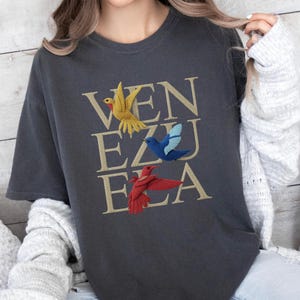 May include: Dark grey t-shirt featuring the word "VENEZUELA" in gold lettering, with embroidered birds in yellow, blue, and red. The t-shirt has a crew neck and short sleeves.
