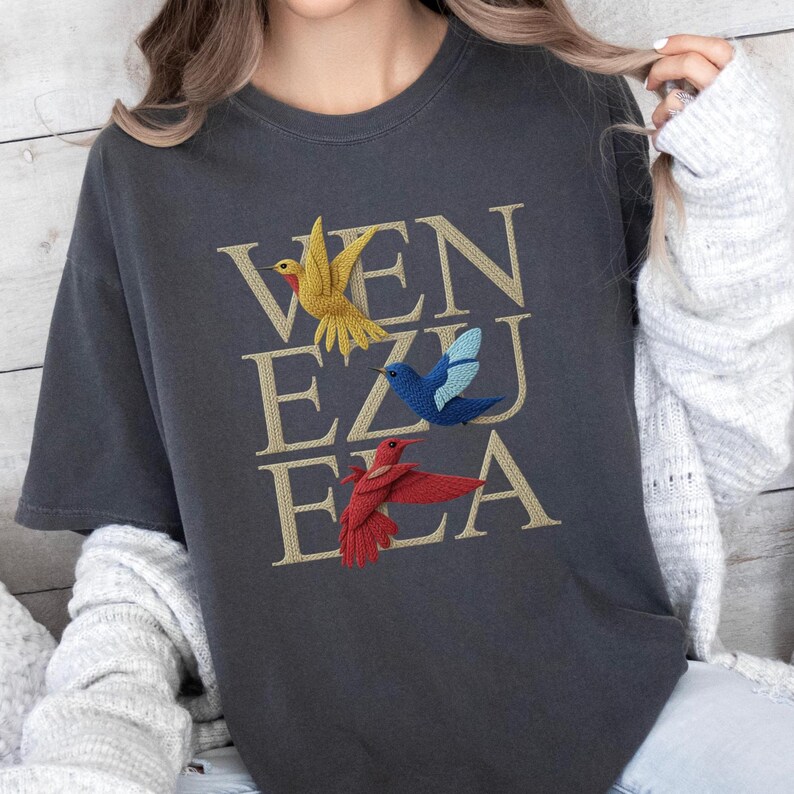 May include: Dark grey t-shirt featuring the word "VENEZUELA" in gold lettering, with embroidered birds in yellow, blue, and red. The t-shirt has a crew neck and short sleeves.