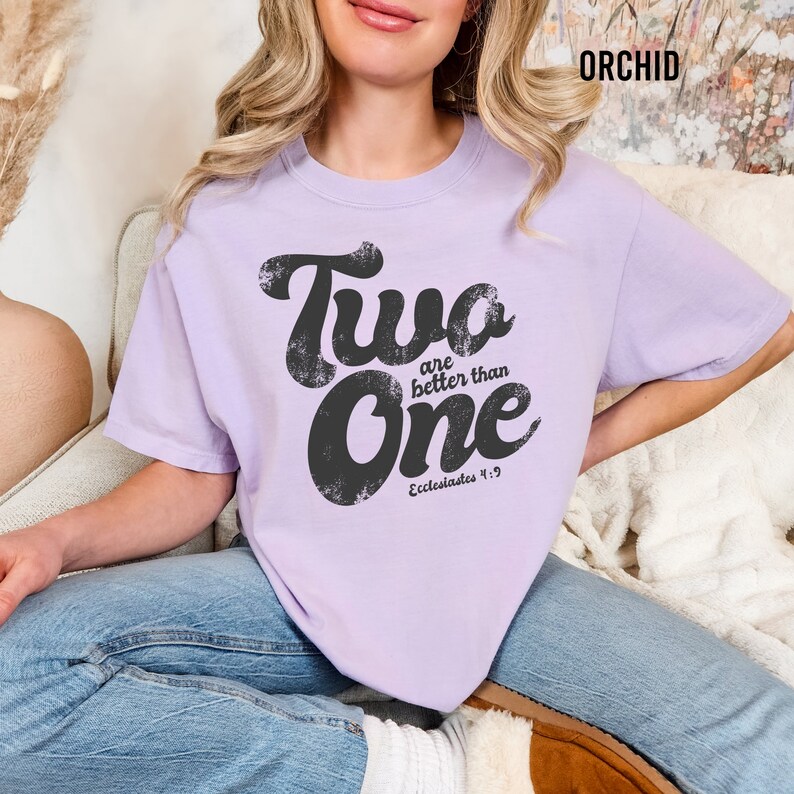 May include: An orchid-colored short-sleeve t-shirt featuring the black distressed text Two are better than One and Ecclesiastes 4:9 printed on the front. The shirt has a crew neckline and a relaxed fit, paired with blue denim jeans.