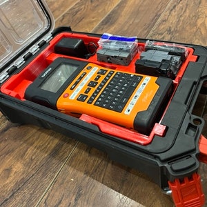 Può includere: Un'etichettatrice nera e arancione con tastiera e display integrati è all'interno di una custodia rigida nera e rossa con un coperchio trasparente. La custodia ha una maniglia e un fermo.