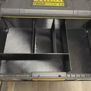 May include: A black toolbox drawer with a yellow trim and the text "TOUGHSYSTEM 2.0" on the top. The drawer is divided into four compartments by black dividers.