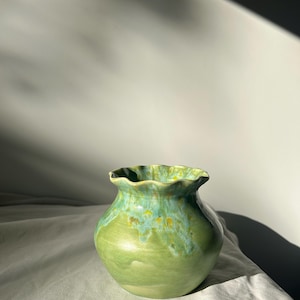 Peut inclure: Un vase en céramique fait à la main avec un bord festonné. Le vase présente un dégradé de nuances de vert, passant d'une teinte plus claire à la base à un turquoise vif au sommet. Des mouchetures jaunes ornent le bord.
