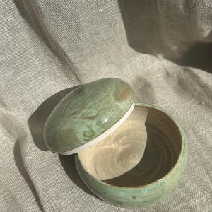 以下が含まれることがあります： 手作りの陶器のボウルと蓋。ボウルと蓋は、マーブル模様のくすんだ緑色です。ボウルの内側は薄いベージュ色です。楕円形の蓋は少し開いています。