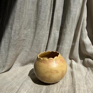 Peut inclure: Un petit vase rond en céramique avec un bord festonné. Le vase a une couleur de base jaune avec des marques mouchetées brun rougeâtre. Le fond est un tissu de ton neutre.
