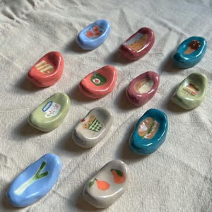 Peut inclure: Ensemble de petits repose-baguettes en céramique. Chaque repose-baguettes est d'une couleur différente et présente un motif unique sur le thème de la nourriture, comme une glace, un avocat et un sandwich. Les repose-baguettes sont disposés sur un tissu beige.