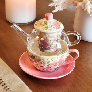 May include: A clear glass teapot with a floral patterned infuser and lid, resting inside a matching floral teacup on a solid pink saucer. The teapot features a curved handle and spout, while the teacup has a pink handle and sits on the saucer.