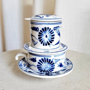 May include: A white and blue ceramic Vietnamese coffee filter set. The set includes a filter, a cup with a handle, a saucer, and a lid. The design features blue floral patterns. The text "Clay & Leaf" is at the bottom.