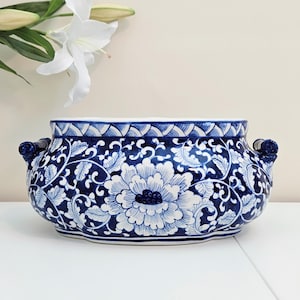 May include: A ceramic bowl featuring a deep blue and white floral pattern with intricate scrolling vines and large peony-like blossoms. The vessel has a wide, rounded body, a scalloped rim with a cross-hatch border, and two small side handles.