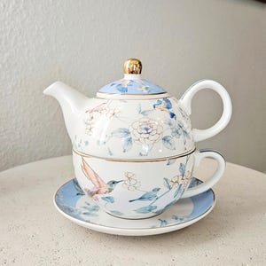 May include: A white porcelain tea set with a blue and gold floral and bird design. The set includes a teapot, teacup, and saucer. The teapot has a gold finial and a curved spout. The teacup and saucer have a blue border.
