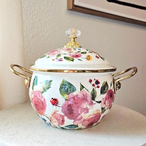 May include: White enamel pot with a floral design featuring pink roses, green leaves, and a ladybug. The pot has gold-colored handles and trim, and a clear knob on the lid.