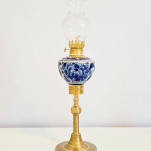 May include: An antique oil lamp with a clear glass chimney and a brass base. The lamp's reservoir is decorated with blue floral patterns on a white background. The lamp is a decorative item.