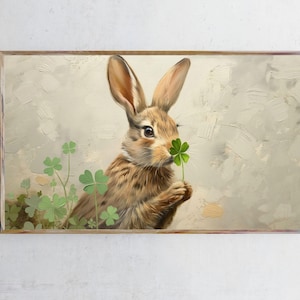 Könnte beinhalten: Gerahmter Kunstdruck mit einem braunen Hasen, der ein grünes vierblättriges Kleeblatt hält. Der Hase hat lange Ohren und ist vor einem neutralen Hintergrund mit grünen Kleeblättern platziert. Das Kunstwerk hat einen Vintage-Malstil.