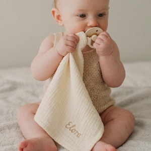 May include: A baby wearing a beige knit romper and holding a cream-colored muslin security blanket with the name "Elora" embroidered in a light brown thread. The baby is also holding a beige pacifier.