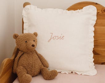 May include: A white square pillow with ruffled edges and the name "Josie" embroidered in a cursive font. A brown teddy bear sits next to the pillow on a pink surface. The pillow is on a wooden bed frame.