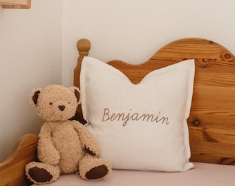 May include: A white pillow with the name "Benjamin" embroidered on it in brown thread. The pillow is on a wooden bed frame with a pink bedspread. A brown teddy bear sits on the bedspread.