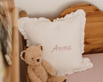 May include: A plush teddy bear sits next to a white pillow with the name "Anna" embroidered in a soft pink script. The pillow has a ruffled edge. The scene is set on a pink surface, near a wooden bed frame.