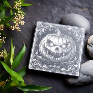 May include: Square black slate coaster with a detailed engraving of a grinning jack-o'-lantern with glowing eyes. The design is framed by an ornate border. The coaster is surrounded by dark stones and greenery, creating a Halloween theme.