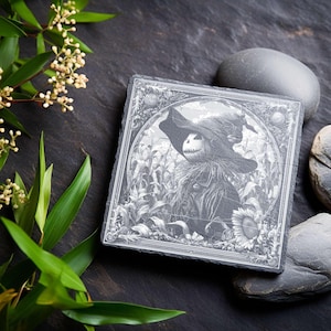 May include: Square slate coaster with a detailed black and white illustration of a scarecrow in a cornfield. The scarecrow has a pumpkin head and is wearing a hat. The coaster is surrounded by a decorative border.