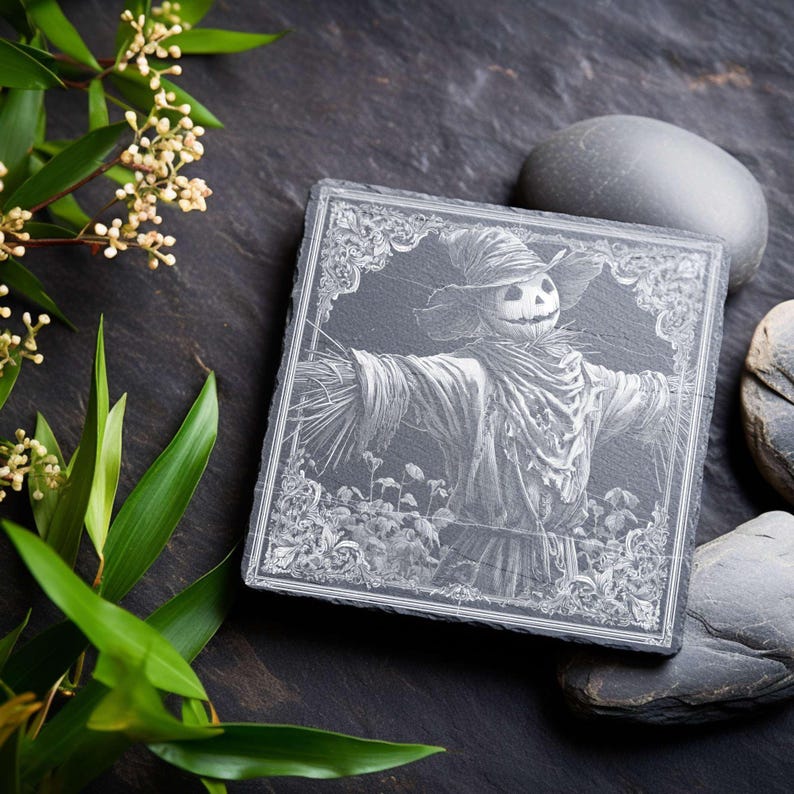 May include: Square, dark gray slate coaster featuring an engraved image of a scarecrow with a smiling face, wearing a hat and a scarf. The coaster has a decorative border and is surrounded by greenery and smooth gray stones.