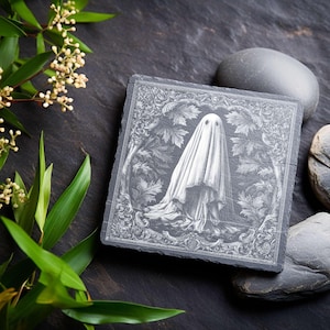 May include: Square slate coaster with a detailed engraving of a ghost figure draped in a white sheet. The ghost is surrounded by a decorative border of leaves and vines. The coaster is dark gray with a textured surface.