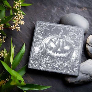 May include: Square slate coaster with a detailed engraving of a jack-o'-lantern with lit candles and foliage. The design is in shades of gray and white, perfect for Halloween decor. The coaster is surrounded by dark gray stones and greenery.