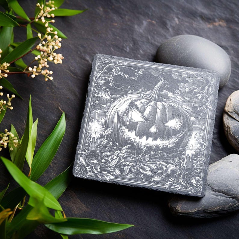 May include: Square slate coaster with a detailed engraving of a jack-o'-lantern with lit candles and foliage. The design is in shades of gray and white, perfect for Halloween decor. The coaster is surrounded by dark gray stones and greenery.