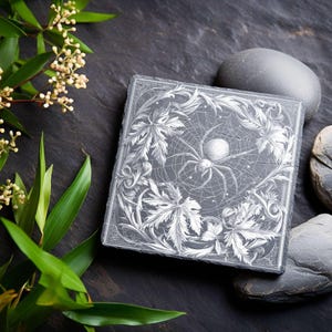 May include: Square, gray slate coaster with a detailed white spider and web design. The coaster is framed by a decorative border of leaves and vines. The coaster is surrounded by smooth gray stones and greenery.