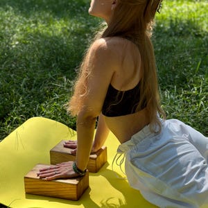 May include: A woman doing yoga outdoors on a yellow mat. She is wearing a black sports bra and white shorts. Two wooden blocks are placed on the mat for support. The background is a green grassy area with trees.