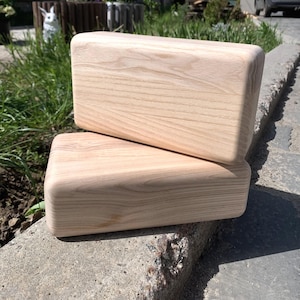 Op de afbeelding: Twee rechthoekige houten blokken met afgeronde randen. De blokken zijn gemaakt van lichtgekleurd hout, mogelijk essen of esdoorn, en hebben een natuurlijk houtnerfpatroon. Ze zijn op elkaar gestapeld, waarbij er één iets schuin staat.