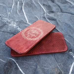 May include: Two red-brown wooden yoga boards. One board features a detailed floral design, while the other has numerous small, evenly spaced metal spikes. The boards are rectangular and rest on a gray stone surface.