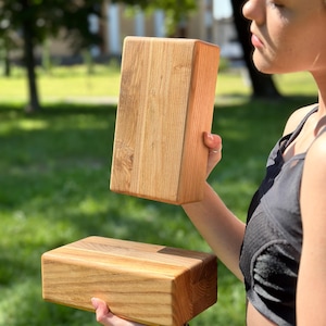 Puede incluir: Dos bloques de yoga de madera, uno sostenido en la mano de una persona y el otro sobre una superficie de hierba verde. Los bloques están hechos de madera natural de color marrón claro con un patrón de veta.