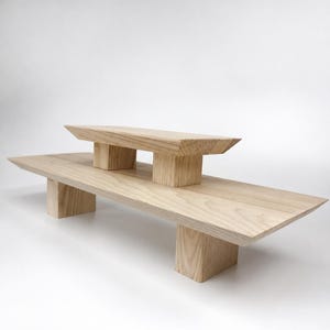 May include: A two-tiered wooden display stand featuring a long, rectangular base platform supported by two block legs and a smaller, elevated rectangular top platform also supported by two block legs, all crafted from light-toned natural wood.