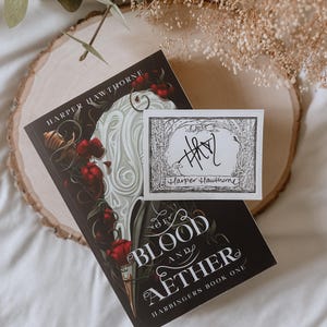 May include: A signed book titled "The Blood and Aether" by Harper Hawthorne. The book cover features a dark background with a white, swirling design and red flowers. The signature is on a white sticker with a black border and a small illustration of a forest.