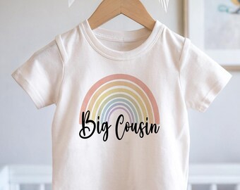 May include: White t-shirt with a rainbow design and the text "Big Cousin" in black.